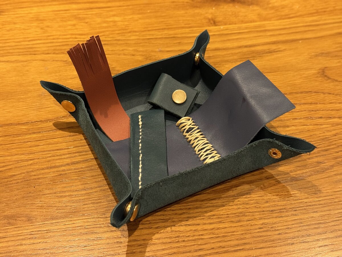 A dark teal leather valet tray on a wooden surface, containing various leather crafting components including rust brown fringed pieces, navy blue leather, and cream coiled strips