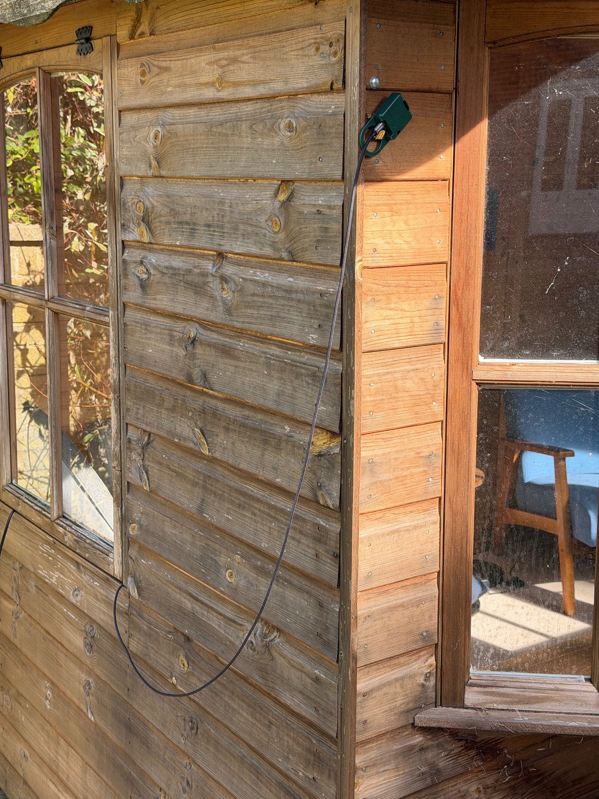 The AudioMoth microphone in its green enclosure, hanging on the wooden wall of the summer house with its USB cable running down the wall.